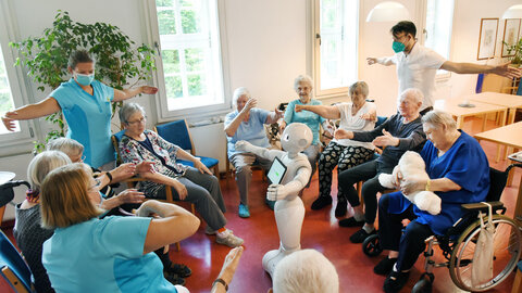A circle of older adults and caregivers, with a robot in the middle demonstrating mobility exercises