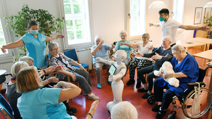 A circle of older adults and caregivers, with a robot in the middle demonstrating mobility exercises