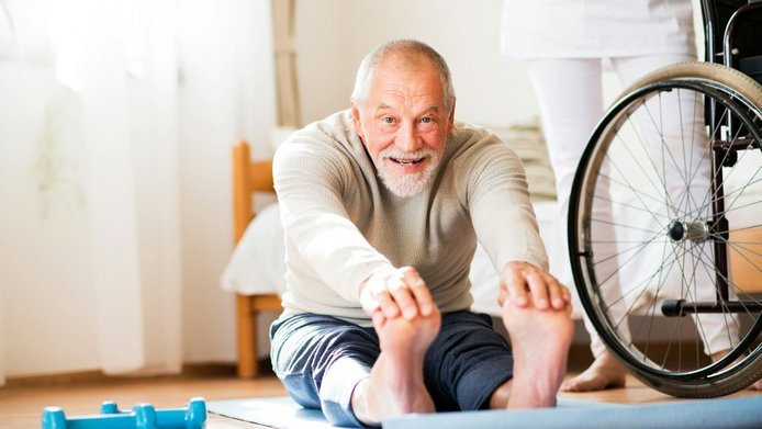 An older man is sitting on a yoga mat on the floor with his legs stretched out, doing stretching exercises; there is a wheelchair next to him