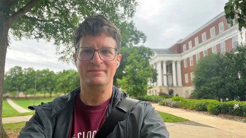 A young researcher wearing glasses and a jacket, standing in front of an old building with a park