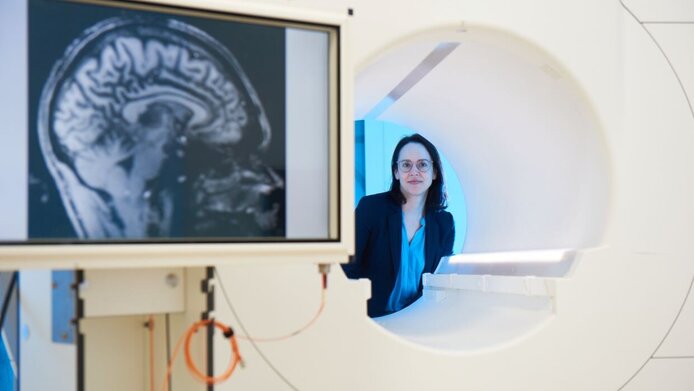 Junge Psychologin im Labor. Sie schaut durch die Öffnung eines Magnetresonanztomografen. Im Vordergrund links ist der Scan eines menschichen Gehirns zu sehen.