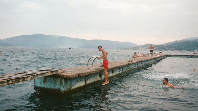 Young man in red swimming trunks climbs out of the sea on a ladder onto an old bathing jetty from the time of the former Yugoslavia.