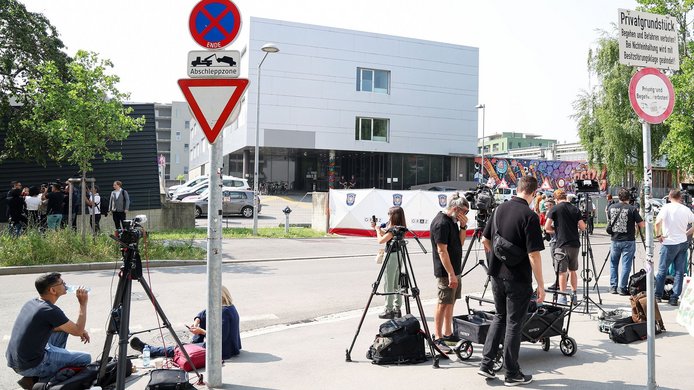 Across from a school building, members of the media are waiting with cameras and video equipment to broadcast live