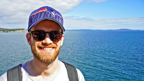 Young man with a full red beard, sunglasses and a cap in front of the sea