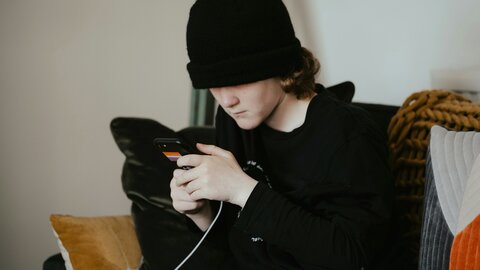 Boy in a black hoodie and black cap sits on the sofa and stares into his smartphone