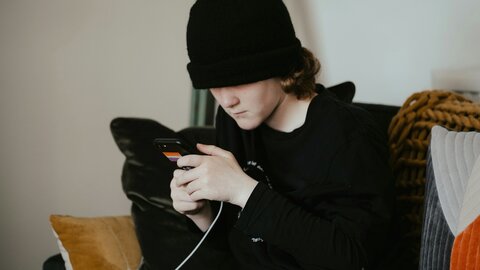 Junge mit schwarzem Kapuzenshirt und schwarzer Mütze sitzt auf dem Sofa und starrt ins Smartphone