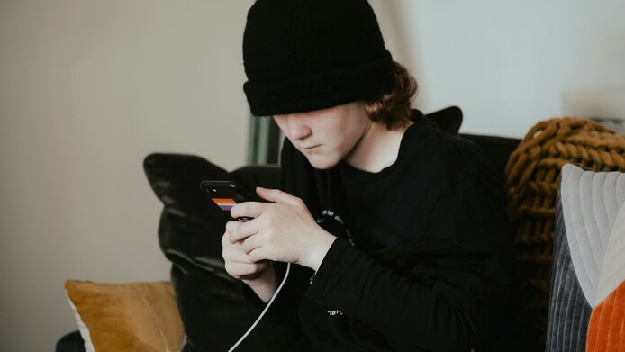 Junge mit schwarzem Kapuzenshirt und schwarzer Mütze sitzt auf dem Sofa und starrt ins Smartphone