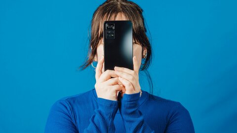 Young woman in a blue sweater in front of a blue background, holding a smartphone in front of her face