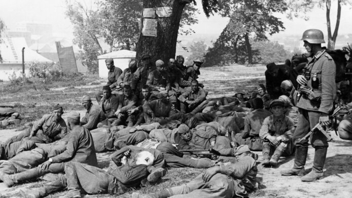 Black-and-white photograph of captured Soviet soldiers during World War II, lying on the ground under a tree and being guarded by a German soldier 