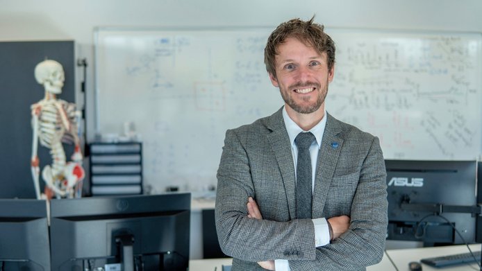 Portrait of a young researcher in a suit, with a workspace featuring computers and a skeleton in the background