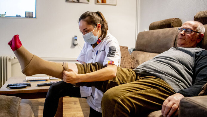 A nurse puts a support stocking on an elderly man's leg in his flat.