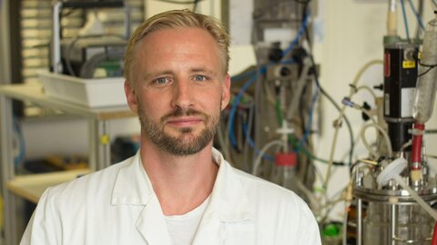 Junger Forscher mit blonder Kurzhaarfrisur und Bart, weißer Kittel, im Labor 