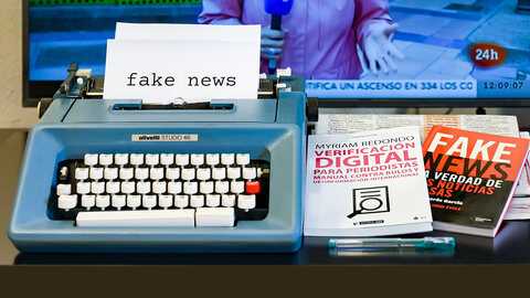 Fake news written on a typewriter, non-fiction books lying next to it, a TV screen in the background