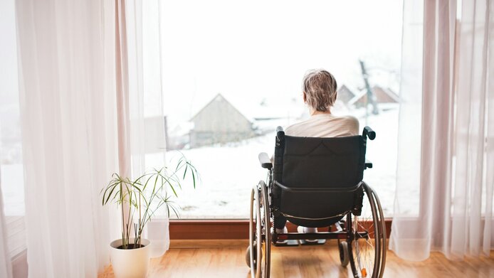 Ältere Frau sitzt im Rollstuhl vor einem großen Fenster und blickt auf eine verschneite Landschaft