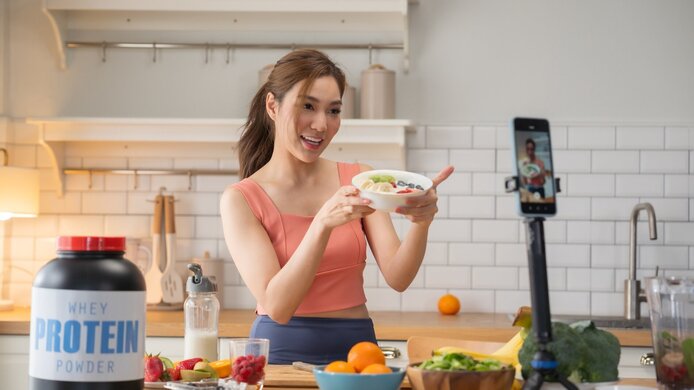 Junge Frau (Influencerin) in der Küche mit Obstteller. Sie erstellt ein Video für die soziale Medien, das ein Nahrungsergänzungsmittel bewirbt