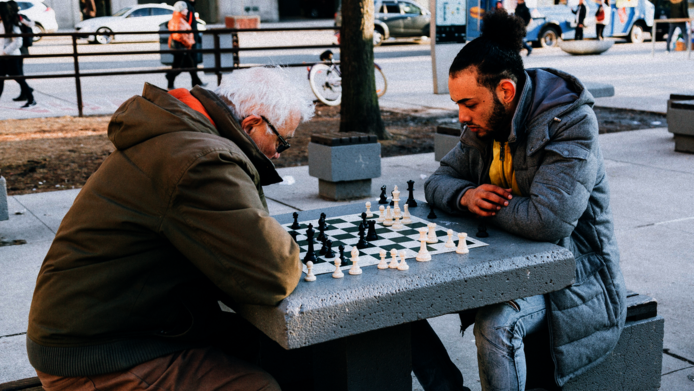 Alter und junger Mann spielen Schach im Freien auf einem Stadtplatz.