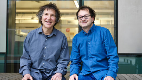 Two male researchers sit on a wooden bench in front of an institute building