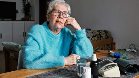 Elderly woman with a buzz-cut hairdo and blue sweater sits thoughtfully at the table on which a blood pressure monitor and medication are lying