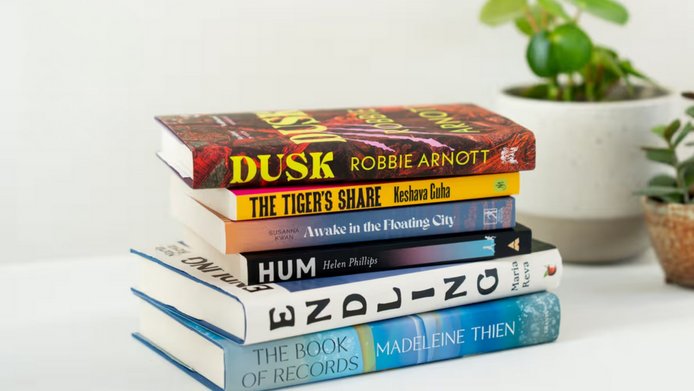 A stack of six books on a table with a small plant in the background