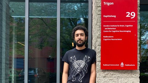 Young neuroscientist with a beard, wearing a T-shirt, leaning against the entrance to his research institute next to a red board.