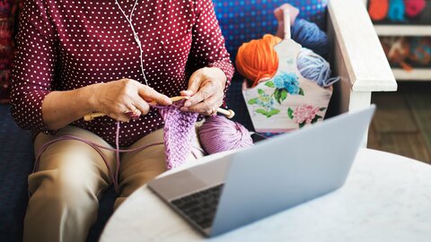 Oberkörper einer älteren Frau, die strickt und vor dem Laptop sitzt