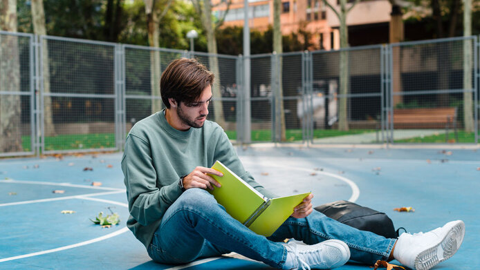 Junger Mann liest in einem Notizbuch, um zu lernen, auf einem Basketballplatz an einer Universität.