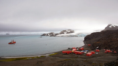 A small container village made up of red huts right on the Antarctic coast. A polar research ship has docked in the sea, with snow and glaciers in the background. 
