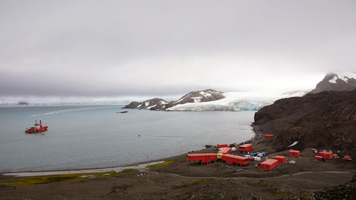 A small container village made up of red huts right on the Antarctic coast. A polar research ship has docked in the sea, with snow and glaciers in the background. 