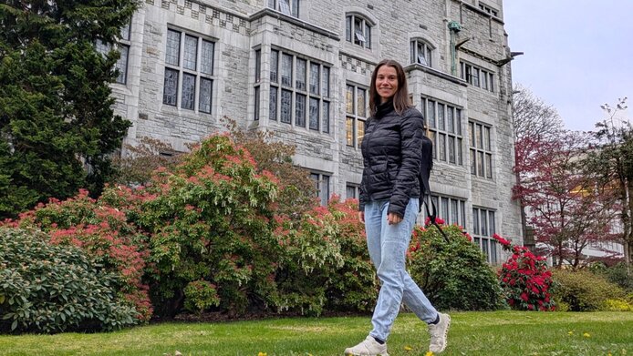 Junge Frau geht an historischem Gebäude einer Universität mit begrüntem Vorplatz vorbei.  