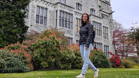 Junge Frau geht an historischem Gebäude einer Universität mit begrüntem Vorplatz vorbei.  