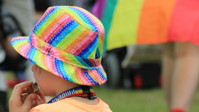 Kleines Kind trägt Hut in Regenbogenfarben während es bei einer Parade in eine Trillerpfeife bläst