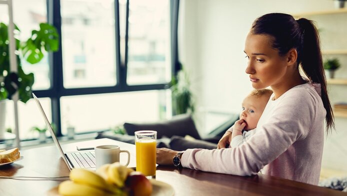 Depressive junge Frau mit Kind auf dem Arm sitzt vor Computer