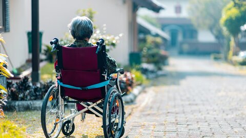 Ältere Frau mit sitzt im Rollstuhl vor ihrem Haus und blickt die Straße hinunter