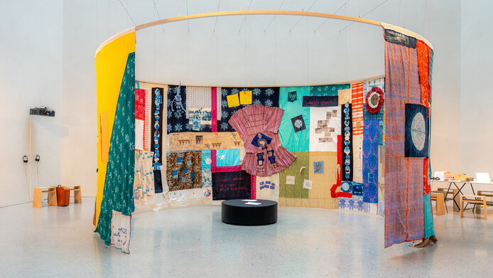 A large quilt made of colorful fabrics and many patterns hangs from the ceiling in a museum.