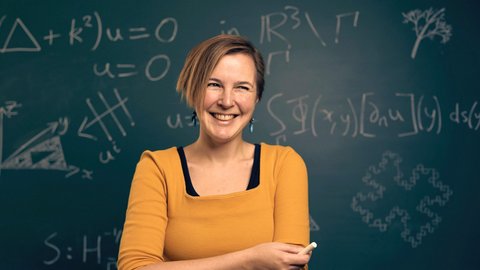 Junge Frau lächelnd mit orangem T-Shirt und Kreide in der Hand vor einer Tafel mit mathematischen Formeln