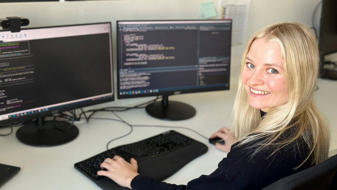 Young woman with long blonde hair in front of two computer screens.